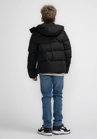 Giacca a vento nera con cappuccio, caratterizzata da un design trapuntato. Indossata sopra una maglietta leggera e abbinata a jeans blu e sneakers nere.