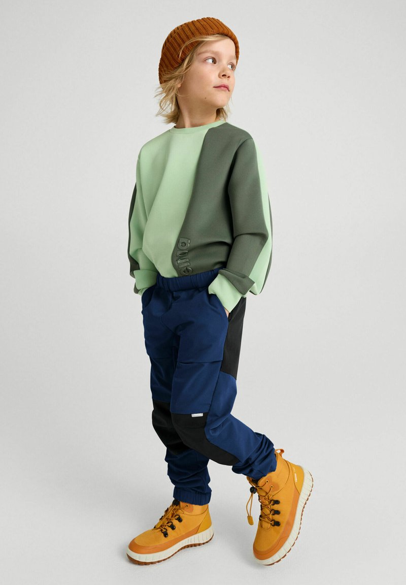 Sudadera en capas verde y gris con botones laterales, combinada con pantalones cargo azules y negros y botas amarillas con suelas blancas.