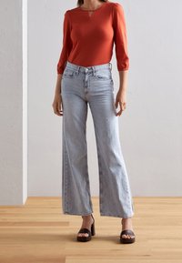 Red long-sleeve top with a keyhole detail, paired with light blue wide-leg jeans and black block-heeled sandals on a wooden floor.