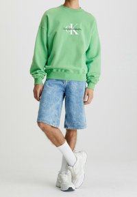 Zelená bavlněná mikina s bílým logem, spárovaná se světle modrými džínovými šortkami. Doplněno bílými ponožkami a botami. Uvolněný střih.