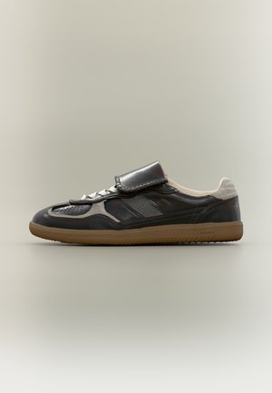 Lavtsiddende sort lædersneaker med beige ruskindsdetaljer, snørebånd foran, delvist dækket af en stor sort læderrem, og brun gummisål.