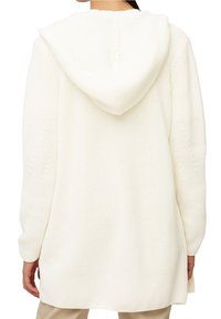 Žena v dlouhém bílém pleteném kardiganu s kapucí, viděná zezadu, s béžovými kalhotami viditelnými dole.