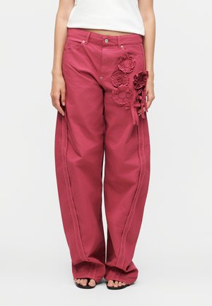 Žena má na sebe voľné hlbokoružové nohavice s kvetinovými textilnými aplikáciami na pravom stehne a čierne sandále s otvorenou špičkou.