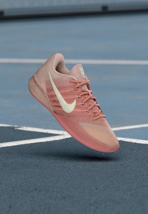 Pink Nike sneaker with white swoosh logo, laces tied, hovering above blue tennis court with white lines.