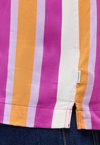 Εγγύς λήψη υφάσματος με πολύχρωμες κάθετες ρίγες σε μωβ, πορτοκαλί και λευκό σε τελείωμα ενδύματος με μια μικρή ετικέτα "Lindbergh" και τζιν υλικό από κάτω.