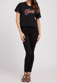 Camiseta negra cropped con el logo "Guess" en rojo, combinada con jeans negros ajustados y sandalias negras de tiras. Tejido suave, mangas cortas y corte relajado.