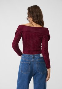 Bordeauxrode trui met blote schouders en kabelgebreide textuur, lange mouwen, gecombineerd met een blauw denim spijkerbroek met hoge taille en achterzakken.