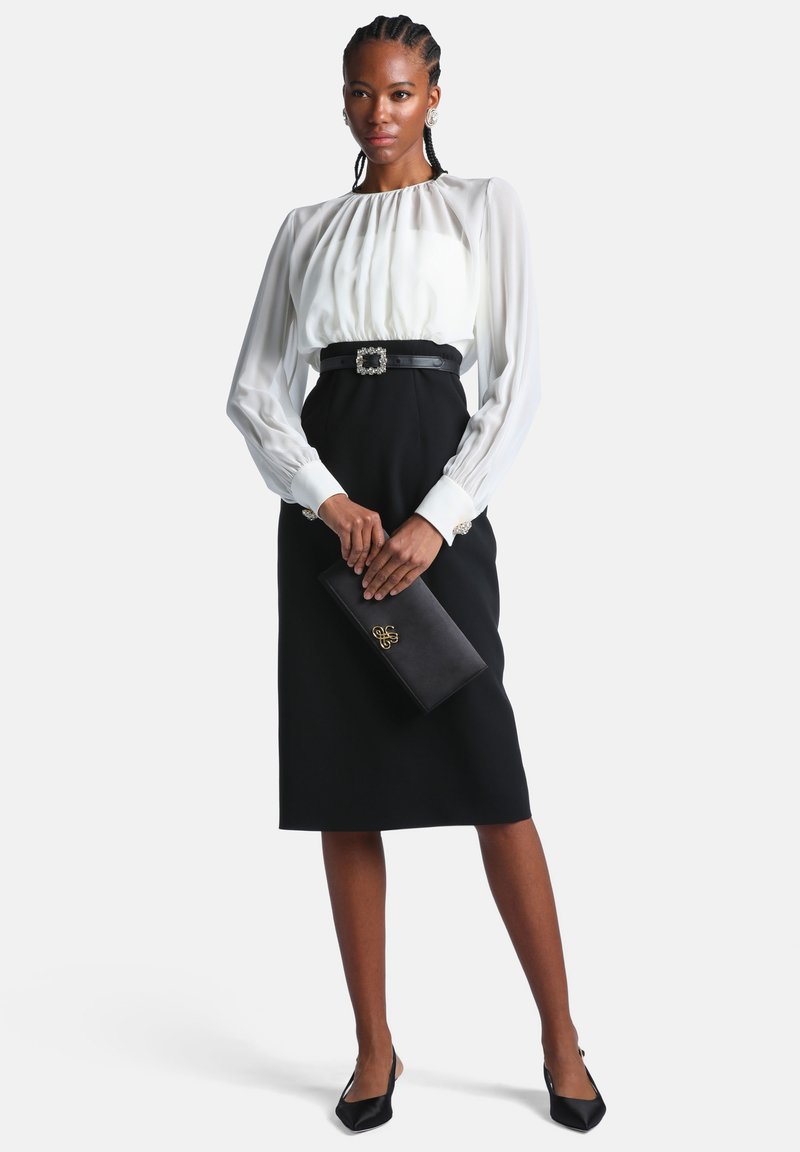 Femme aux cheveux tressés portant un chemisier blanc à manches longues, une jupe noire taille haute, une ceinture noire avec boucle, tenant une pochette noire.