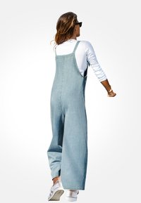 Jasnoniebieskie dżinsowe ogrodniczki z szerokimi nogawkami i regulowanymi ramiączkami, w połączeniu z białą bluzką z długim rękawem i białymi sneakersami.