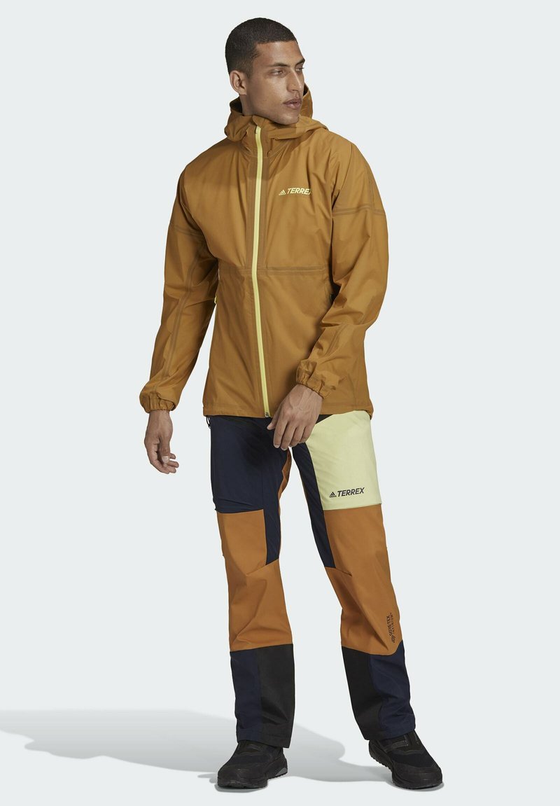 Chaqueta impermeable marrón con cremallera amarilla, combinada con pantalones en bloques de color marrón, azul marino y amarillo claro, que presentan un logo en el muslo.