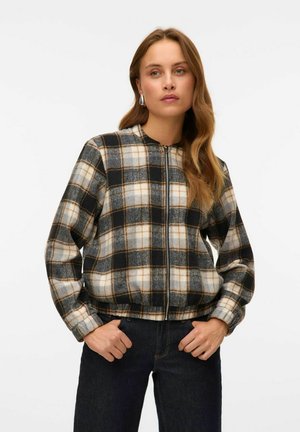 Kvinna iklädd en zip-up, svart, vit och brun rutig jacka med elastisk nederkant och mörka jeans, stående med händerna i fickorna mot en vit bakgrund.