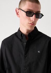 Black button-up shirt with a collared neck, featuring a small embroidered logo. Smooth texture and regular fit, complemented by black sunglasses.