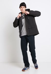 Chaqueta exterior negra con capucha, sudadera gris debajo, pantalones oscuros y zapatillas multicolores. Tejido suave con bolsillos visibles.