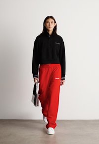 Czarny krótki sweatshirt z zamkiem, czerwone szerokie spodnie dresowe z białym brandingiem, w połączeniu z białymi sneakersami. Prosta, sportowa stylizacja.