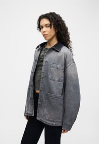 Γυναίκα με σγουρά μαλλιά που φοράει μια φαρδιά ξεθωριασμένη γκρι denim τζάκετ πάνω από ένα σκουρόχρωμο κοντό μπλουζάκι και μαύρο παντελόνι, στέκεται μπροστά σε ένα απλό φόντο.