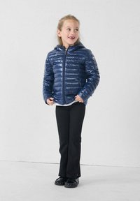 Marine blauwe pufferjack met een gladde, glanzende textuur, ritsluiting en capuchon, gedragen over een wit shirt en zwarte broek. Staand tegen een lichte achtergrond.