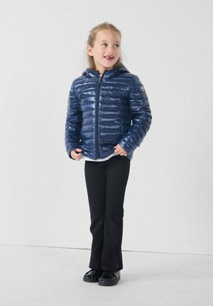 Giacca puffy blu marina con una texture liscia e lucida, chiusura con zip e cappuccio, indossata sopra una camicia bianca e pantaloni neri. In piedi contro uno sfondo chiaro.
