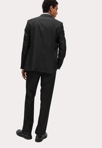 Czarny garnitur z dopasowanym krojem, gładkim materiałem i jednym rozcięciem z tyłu; w komplecie z czarnymi eleganckimi butami. Żakiet ma klapę typu notch.