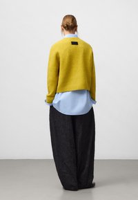 Geel wollen trui, gelaagd over een lichtblauw shirt, gecombineerd met zwarte gestructureerde wijde broekspijpen. Heeft een zwart label aan de achterkant.