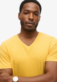 T-shirt giallo in cotone con scollo a V, maniche corte, tessuto leggero e vestibilità rilassata, indossato da una persona con le braccia incrociate.