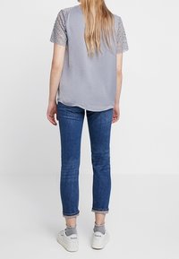 Top gris de manga corta con detalles de encaje en las mangas, combinado con jeans ajustados azules y zapatillas blancas. El aspecto general es informal.