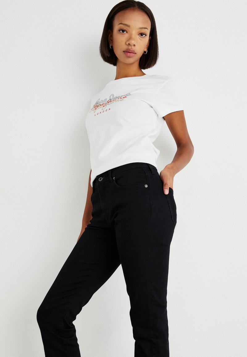 Jeans negros combinados con una camiseta blanca que presenta un texto de logo en rojo y plata. El corte es slim con bolsillos delanteros y un diseño clásico.