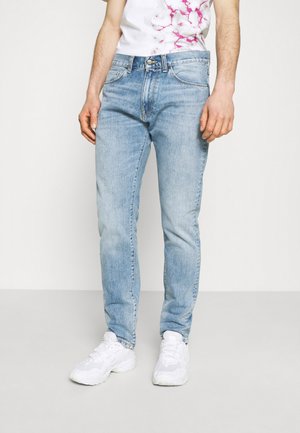 Jeans de ganga azul claro com um corte slim, apresentando bolsos frontais e fecho de zipper. O tecido tem uma textura suave e um ligeiro desbotamento.