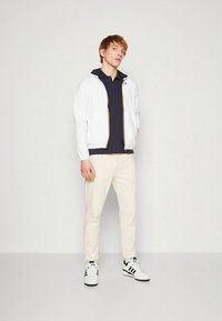 Biela bunda na zips so zlatým zipsom, tmavomodré polokošeľa, béžové nohavice, čierno-biele tenisky, stojaci na neutrálnej pozadí.