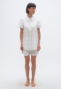 Femme aux cheveux courts et foncés portant une chemise blanche à boutons avec des bords festonnés et un short blanc assorti, debout pieds nus sur un fond uni.