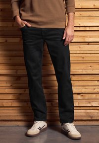 Persona con pantalones negros, zapatillas blancas y una camiseta de manga larga marrón, de pie contra una pared de listones de madera con una mano en el bolsillo.