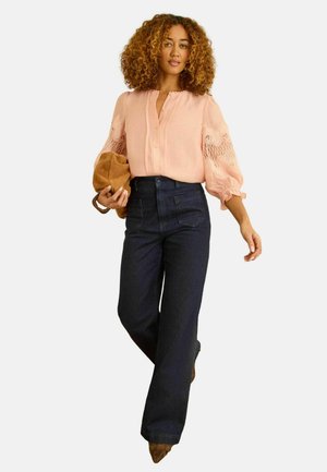 Sieviete ar cirtainiem matiem rozā mežģīņu blūzē un tumšās platām kāju džinsās, tur rokās brūnu zamšādas somu, ejot uz priekšu.