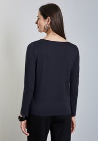 Pitkähihaisella tummansinisellä paidalla, jossa on leveä pääntie, sileä tekstuuri ja minimaliset saumat, yhdistettynä mustiin housuihin ja paksuun rannekoruun.