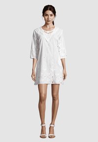 Vestido túnica de algodón blanco bordado con mangas tres cuartos, detalles de encaje y patrones florales. Presenta cortes decorativos y un ajuste relajado.
