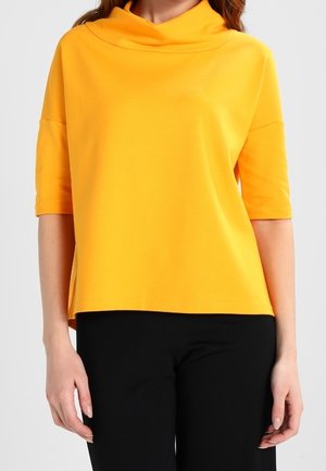 Woman wearing a bright yellow elbow-sleeve top with a draped neckline and black pants, standing against a plain background.