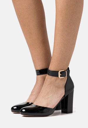 Jambes portant des chaussures à talons carrés en cuir verni noir brillant avec des brides à la cheville et des boucles dorées sur un fond clair.