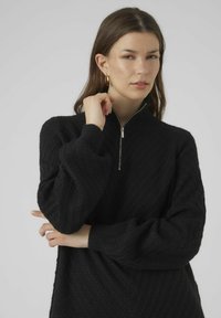 Maglione zip-up nero con un motivo a maglia testurizzato, colletto alto e maniche ampie. Il materiale sembra morbido e leggermente peloso.