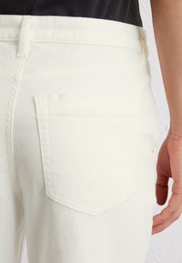 Jeans de denim blanco con un bolsillo trasero con costuras finas y una ligera textura. La tela parece suave con un diseño ajustado.