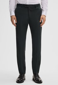 Op maat gemaakte broeken van donkergroene stof, met een platte voorkant, riemlussen en zijzakken. Gecombineerd met zwarte nette schoenen.