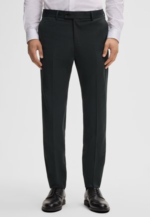 Pantaloni su misura in tessuto verde scuro, con fronte piatto, passanti per la cintura e tasche laterali. Abbinati a scarpe eleganti nere.