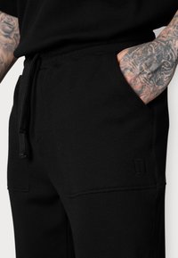 Schwarze Ripp-Jogginghose mit einem elastischen Bund und Zugband. Sie verfügt über Seitentaschen und ein dezentes Logo-Detail nahe dem Saum.