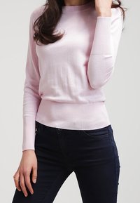 Maglione lunghezza maniche rosa chiaro con scollatura rotonda e orlo a coste. Abbinato a jeans scuri, che evidenziano una texture liscia e un design aderente.