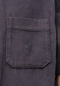 Μεγάλη τσέπη από βελούδινο ύφασμα σκούρου γκρι με κάθετες αυλακώσεις, ραμμένη σε φόντο από ίδιο ύφασμα, εμφανίζοντας μέρος ενός βραχίονα στη δεξιά πλευρά.