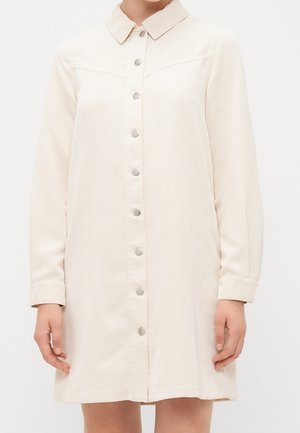 Beiges Cordhemdkleid mit langen Ärmeln, Druckknöpfen vorne und klassischem Kragen, getragen von einer stehenden Person.