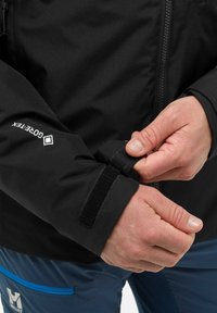 Schwarze wasserdichte Jacke mit verstellbaren Klettverschlussärmeln, mit einer glatten Textur und Logodetail am Ärmel. Die Hose ist blau mit blauen Akzenten.