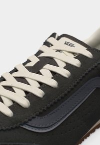 Κοντινό πλάνο μαύρου αθλητικού παπουτσιού Vans με λευκά κορδόνια και χαρακτηριστική πλευρική ρίγα, αναδεικνύοντας το λογότυπο στη γλώσσα και το υφή σουέτ υλικό.