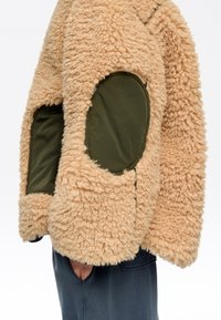 Chaqueta beige de forro polar rizado con grandes parches ovalados verde oliva en las mangas y bolsillo, llevada por una persona con la mano en el bolsillo de pantalones oscuros.