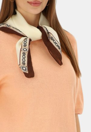 Kobieta w brzoskwiniowym swetrze z krótkim rękawem i kremowo-brązowym jedwabnym szaliku zawiązanym wokół szyi.