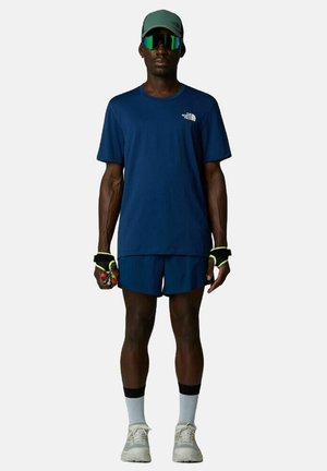 Camiseta atlética de la marina y pantalones cortos, tela ligera, detalles reflectantes, gorra verde, gafas de sol grandes, calcetines blancos y zapatillas deportivas.