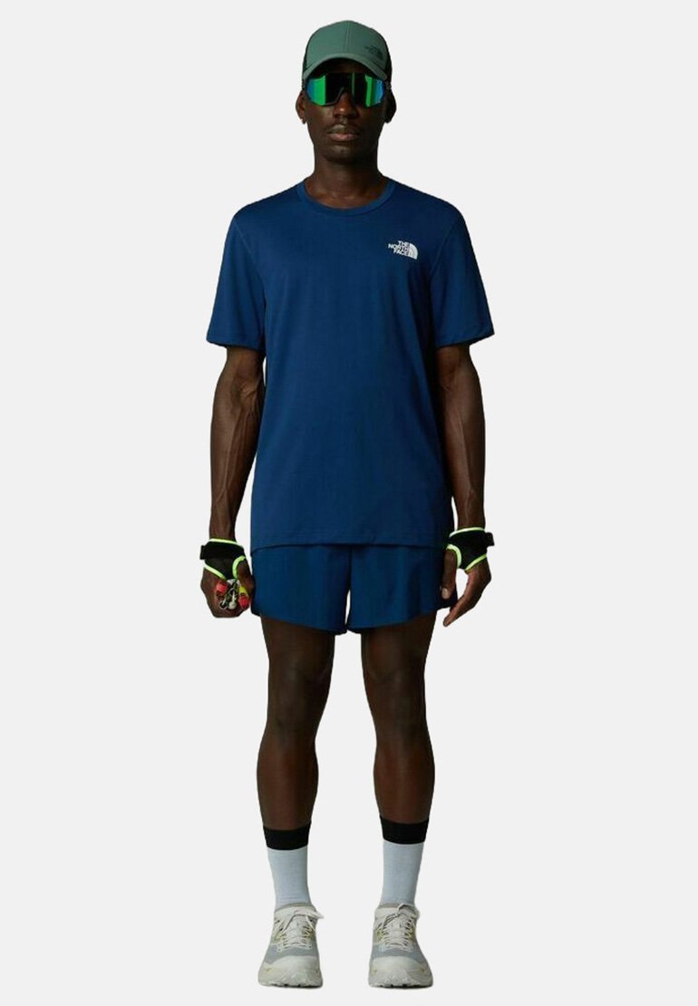 Marinefarbene Sport-T-Shirt und Shorts, leichter Stoff, reflektierende Details, grüner Hut, übergroße Sonnenbrille, weiße Socken und Sportschuhe.
