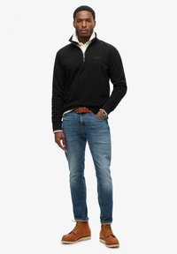 Pullover in pile nero con zip, colletto khaki, jeans blu e stivali beige stringati. Presenta un logo sul petto e una vestibilità classica.